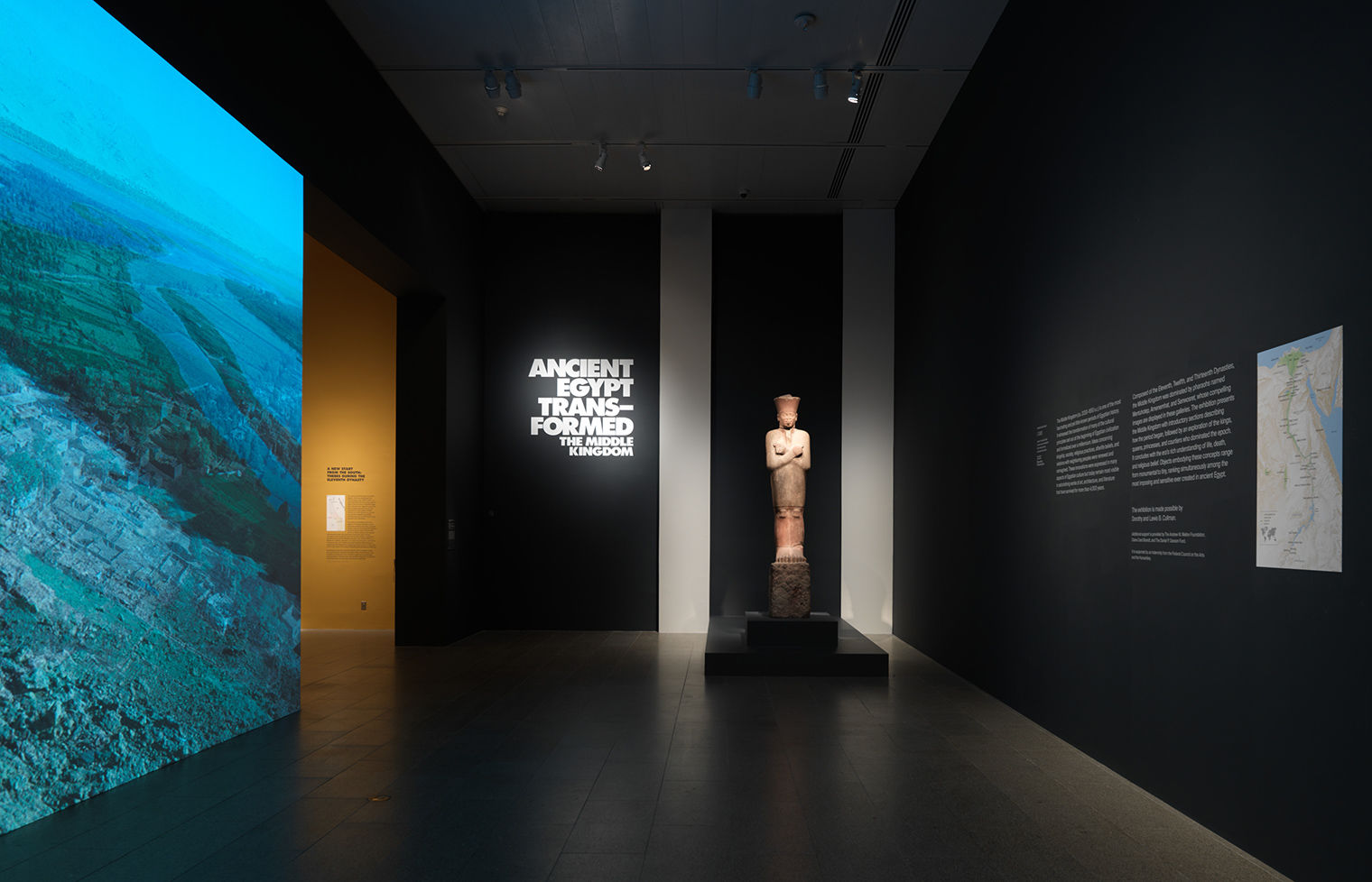 The entrance to the exhibition, Ancient Egypt Transformed, with a projection of an Egyptian landscape to the left and a statue of a king wearing a knee-length white robe and a red crown with a flat top straight ahead.