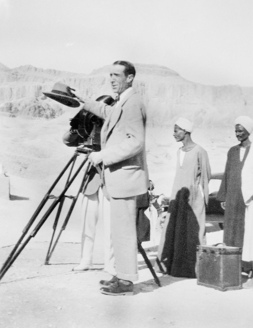 In the desert with cliffs in the background, an archaeologist in a suit, gesturing with his hat while another man takes photo with a camera on a tripod and two other men in turbans and long robes watch.