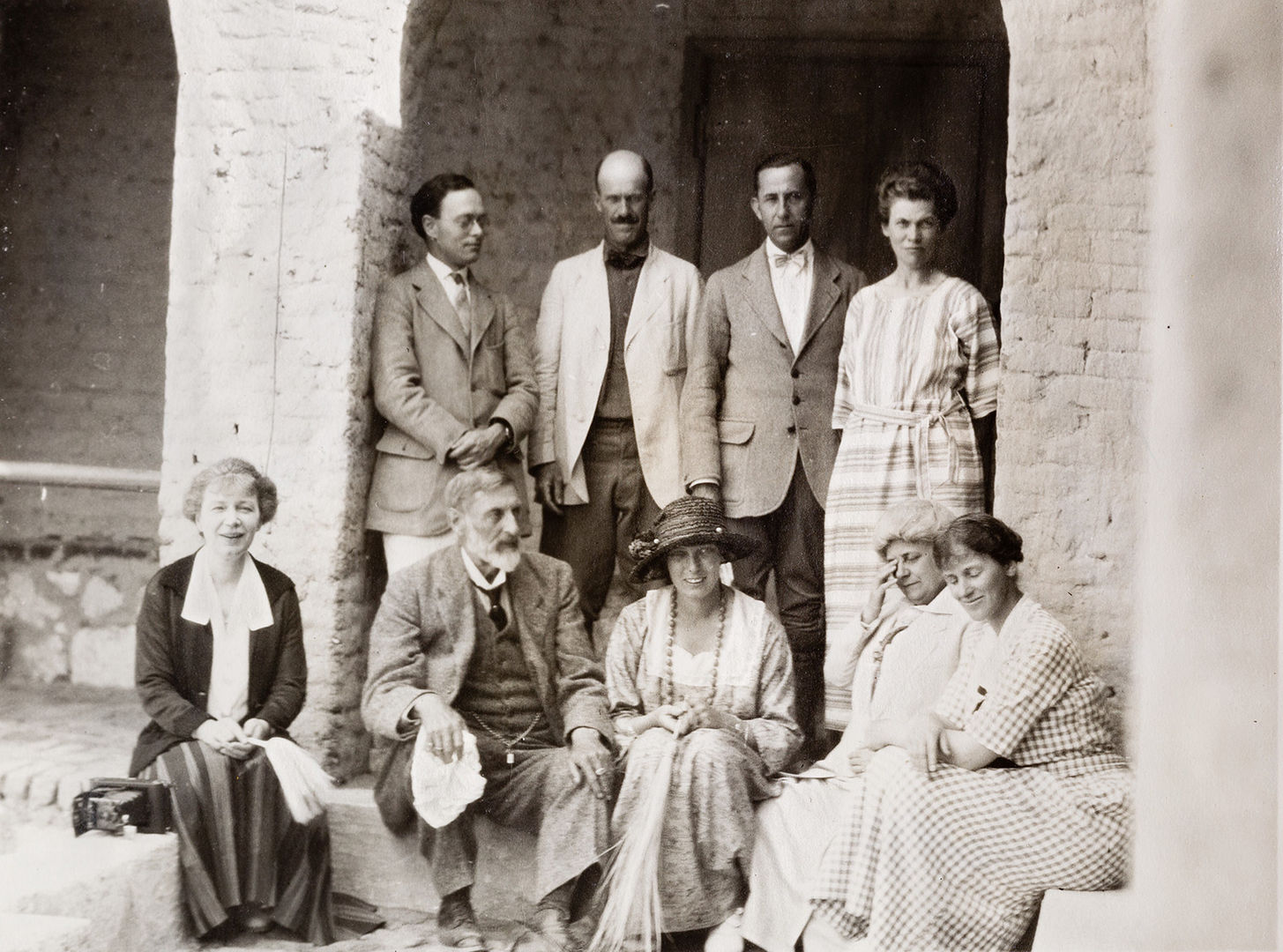 A group of nine people pose in the archway of a house, with three men and one woman in the back and one man and four women in the front. 