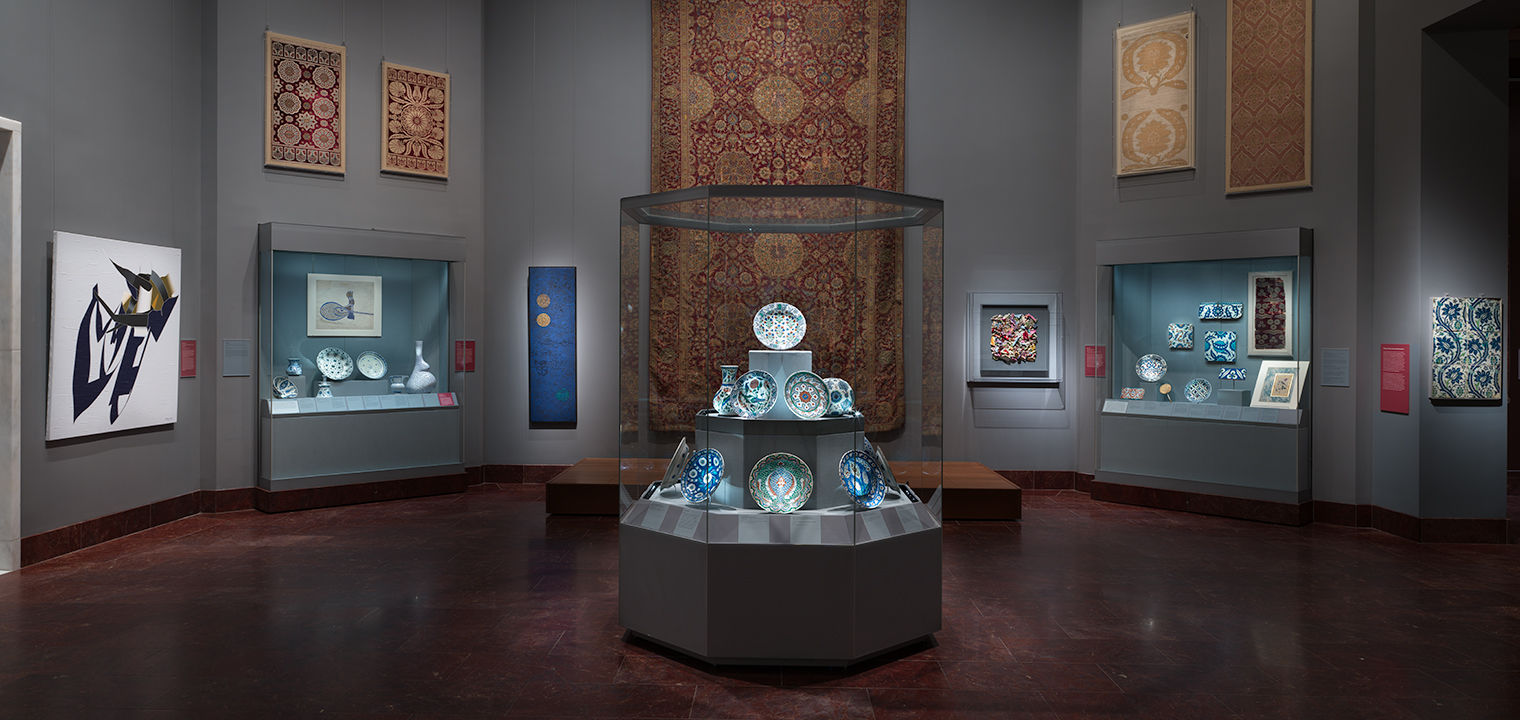 Gallery view of the installation, Dialogues: Modern Artists and the Ottoman Past, in the Koç Family Galleries - Carpets, Textiles and the Greater Ottoman World and Arts of the Ottoman Court (14th–20th centuries) depicting a central glass case with Ottoman ceramics in the foreground, and wall cases, carpets and textiles, and modern artworks on the walls in the background