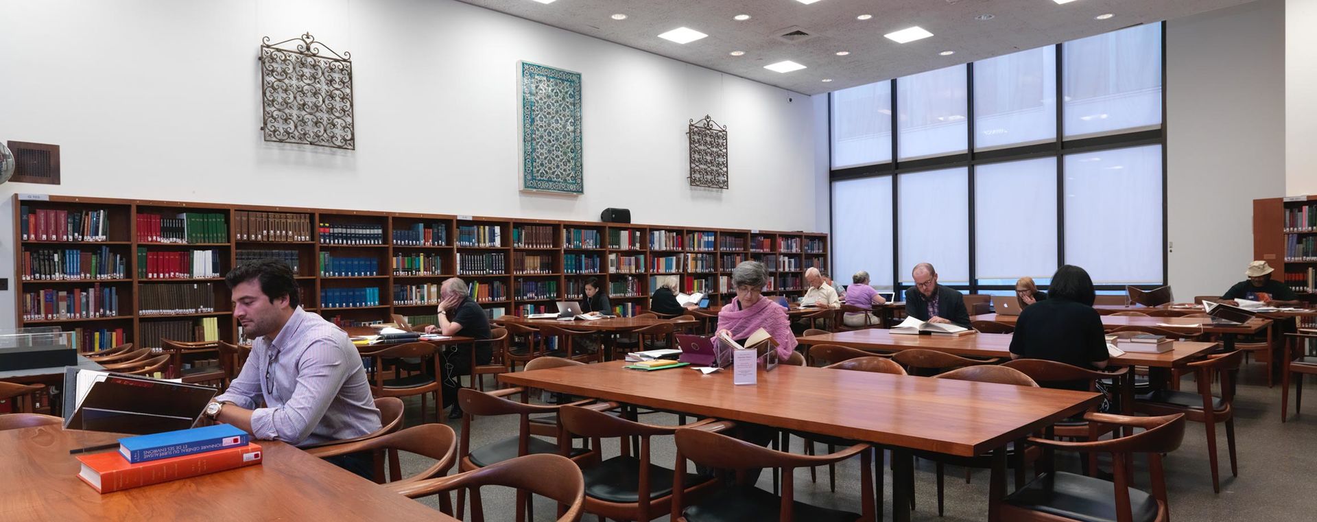 A large, brightly lit room in a library lined with shelves of books; the center has long rectangular wooden tables and chairs where researchers work.