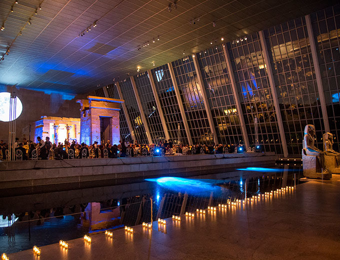 Large glass-enclosed gallery at night decorated with lit with candles and dramatic lighting for a party; in the center of the gallery is an ancient Egyptian sandstone temple 