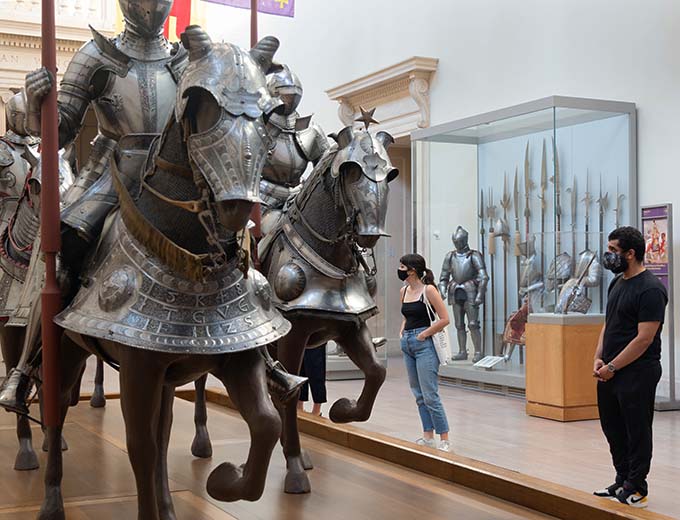 A man and a woman wearing face masks look at armor on display in the Equestrian Court at The Met Fifth Avenue.