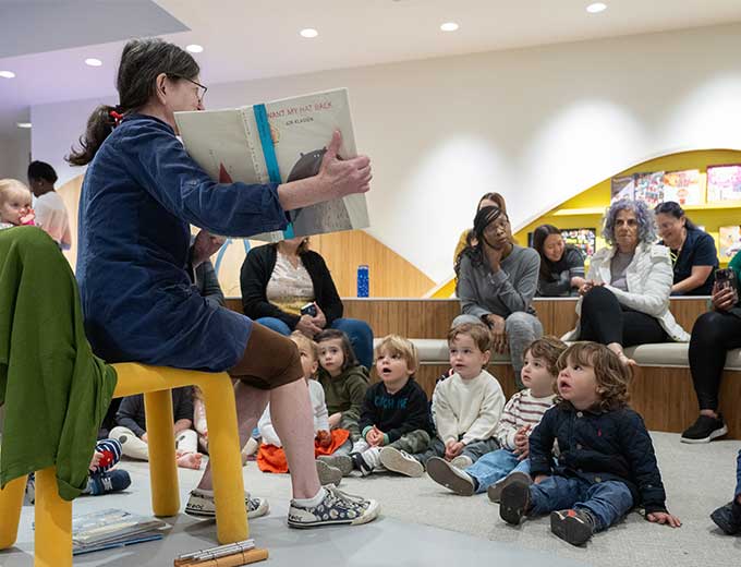A woman sits on a yellow chair. See is holding open a picture book, and a group of toddlers seated before her on the floor are listening to her reading the story.