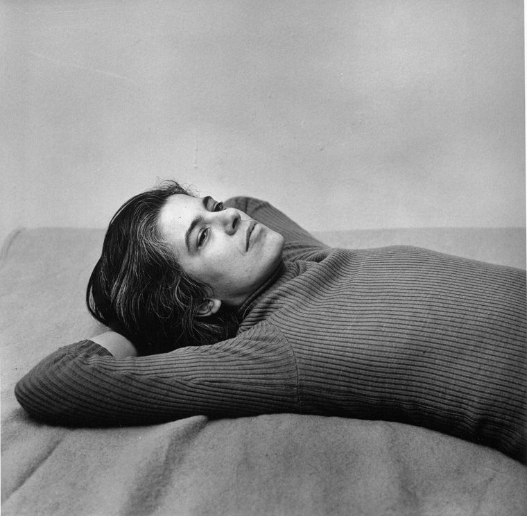 Black-and-white square photograph of Susan Sontag wearing a turtleneck sweater and lying on her back, photographed by Peter Hujar