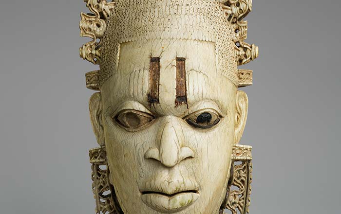 Image of an ivory pendant mask portraying the Queen Mother, Idia, mother to the king of Benin. The mask has inlaid metal and carved scarification marks on the forehead. She is wearing bands of coral beads below the chin. In the openwork tiara and collar are carved stylized mudfish and the bearded faces of Portuguese