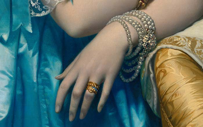 Close-up detail of a woman's hand resting against a yellow chair. She is wearing gold rings and a multi-stranded pearl bracelet. 