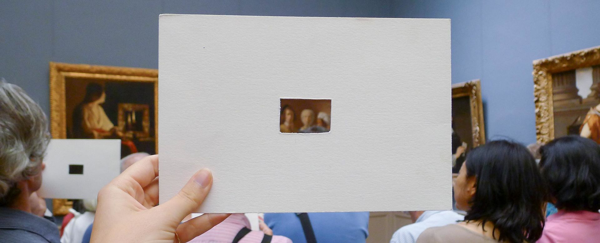 Visitors in a paintings gallery during a class; a visitor is viewing a portion of a painting through a small rectangle cut into a piece of white cardboard