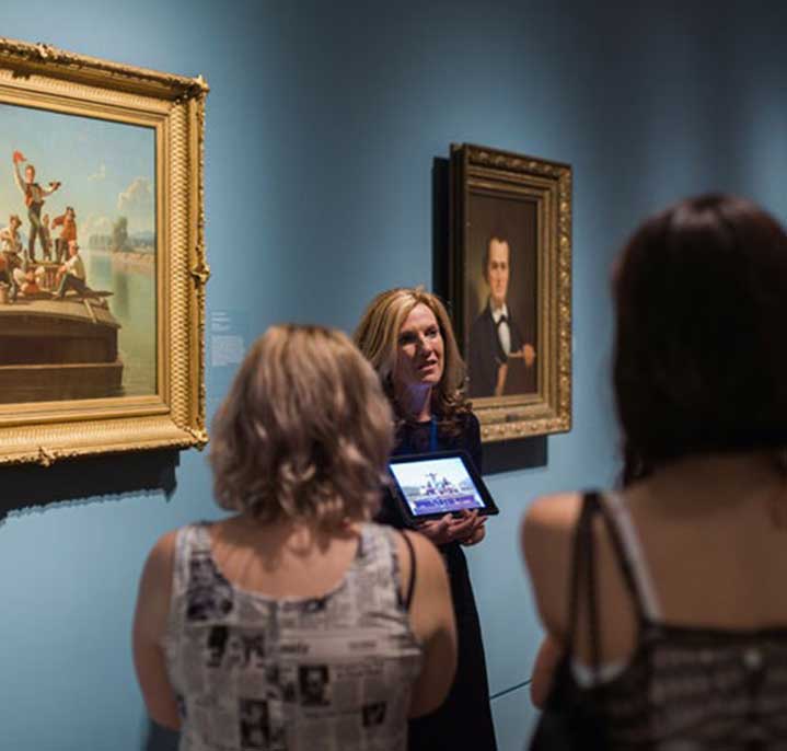 A Museum tour guide addresses a small group of visitors. She is holding an iPad and standing in front of an oil painting in a gallery with blue walls.