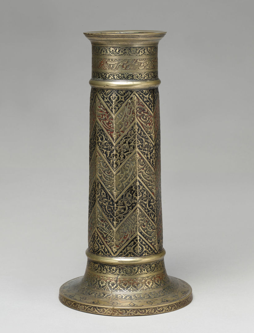 A brass candle stick engraved wtih a zigzag pattern and decorated in inlaid black and red pigment