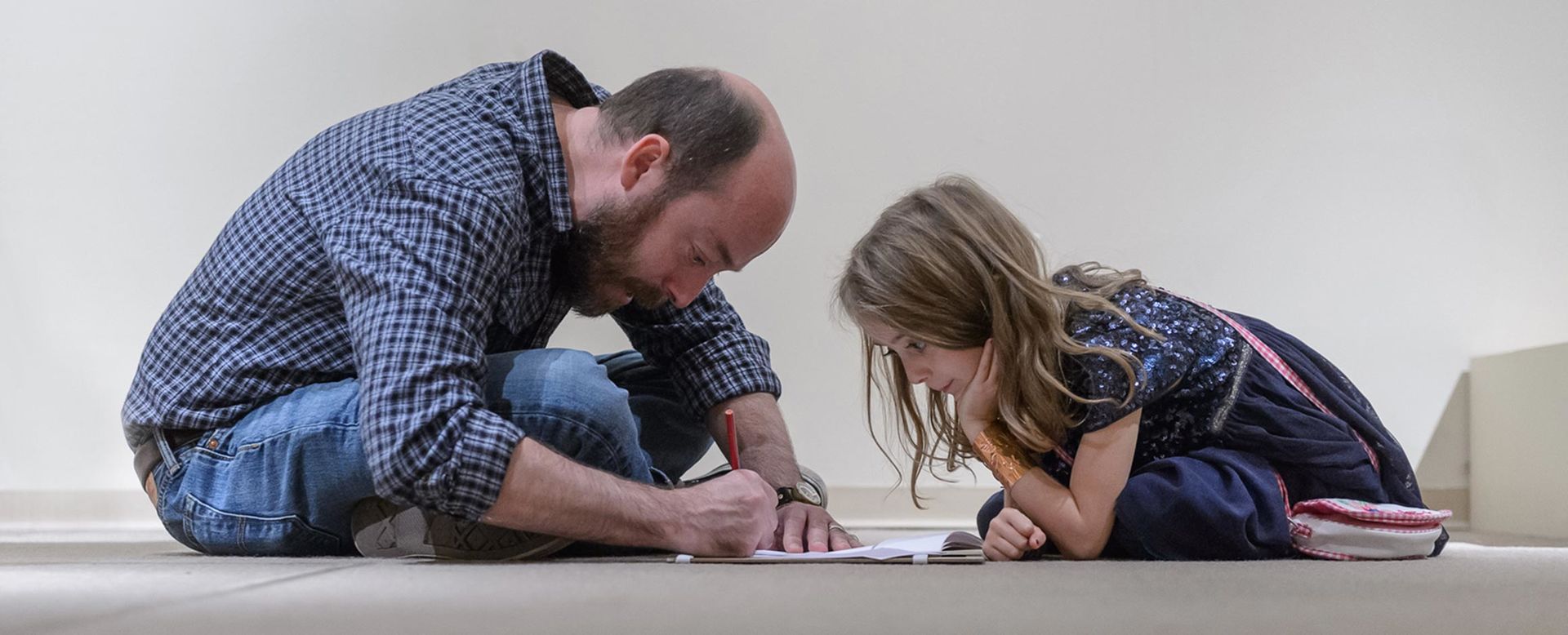 A young girl sits watching a bearded man sketch in the middle of the gallery floor