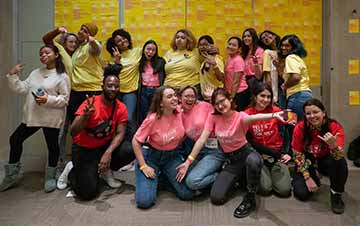 A group of teens are waving and making fun faces at the camera. They are wearing pink and yellow t-shirts, and look very happy.
