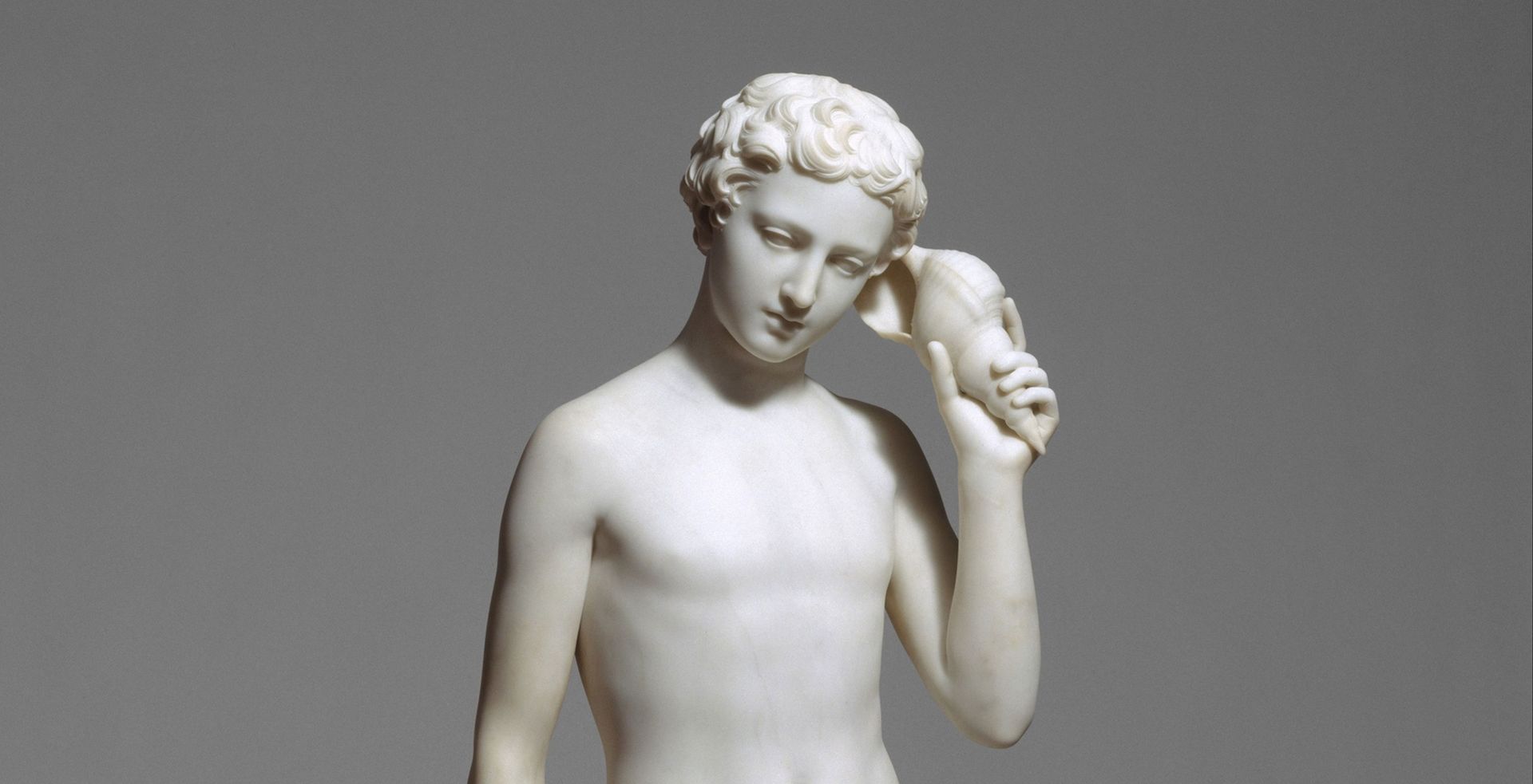 A white marble carving of a muscular shirtless boy with curly hair holding a conch shell up to his ear and listening.