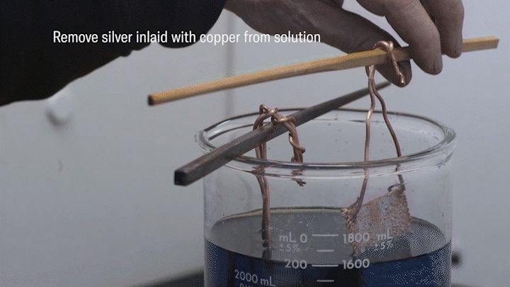 A video animation of an artisan detaching the power source, and then removing silver inlaid with copper solution. The artisan then removes excess plated copper with pliers before smoothing the surface with a polishing stone.
