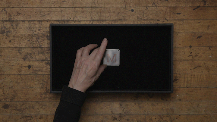 A video animation of an artisan placing a finished work of small silver plate with an inlay design of a copy letter M against a black felt background.