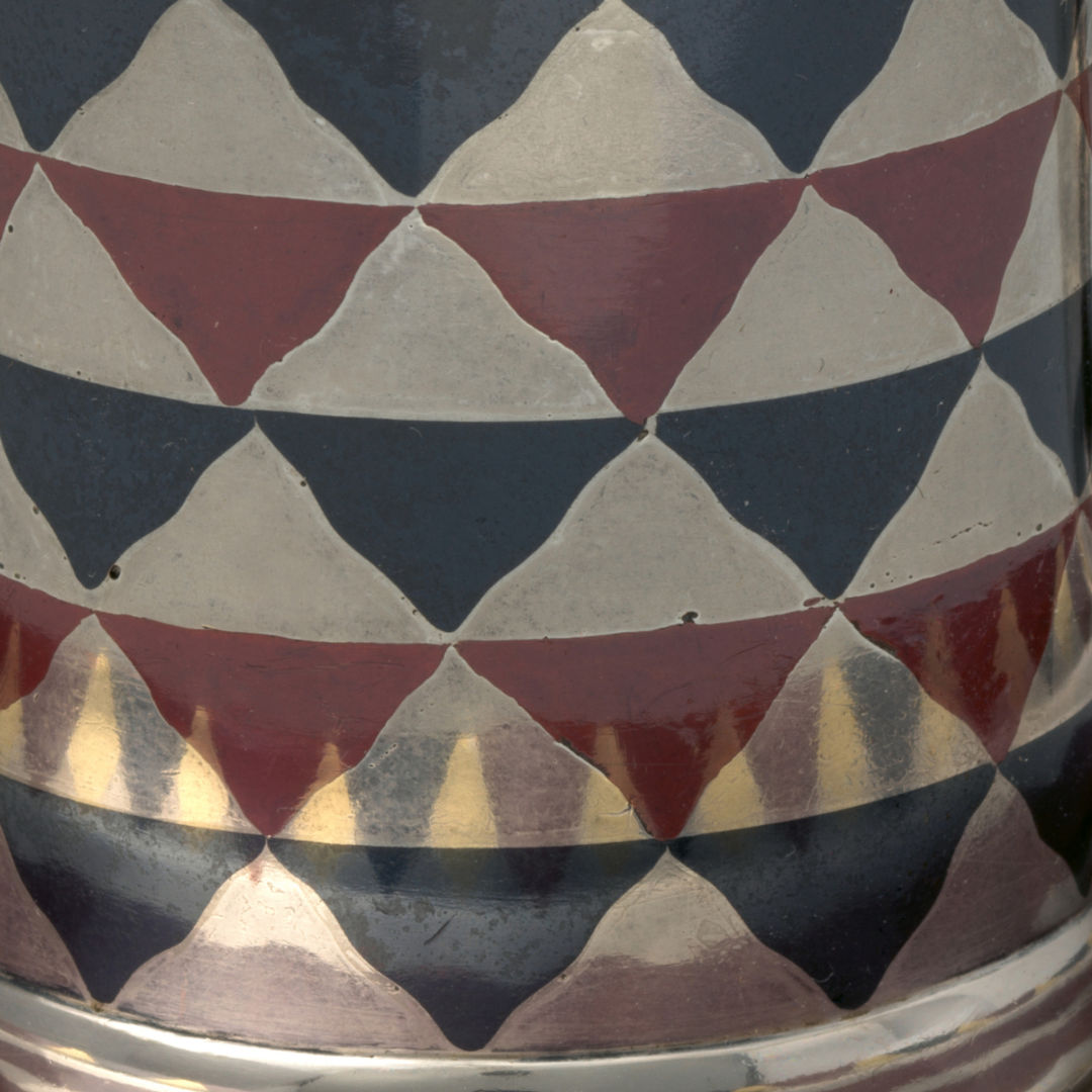 A close up view of silver, blue, and maroon cylindrical cup. The surface of the cup is reflective and is adorned with an inlay design of a checkerboard pattern of wavy triangle shapes.