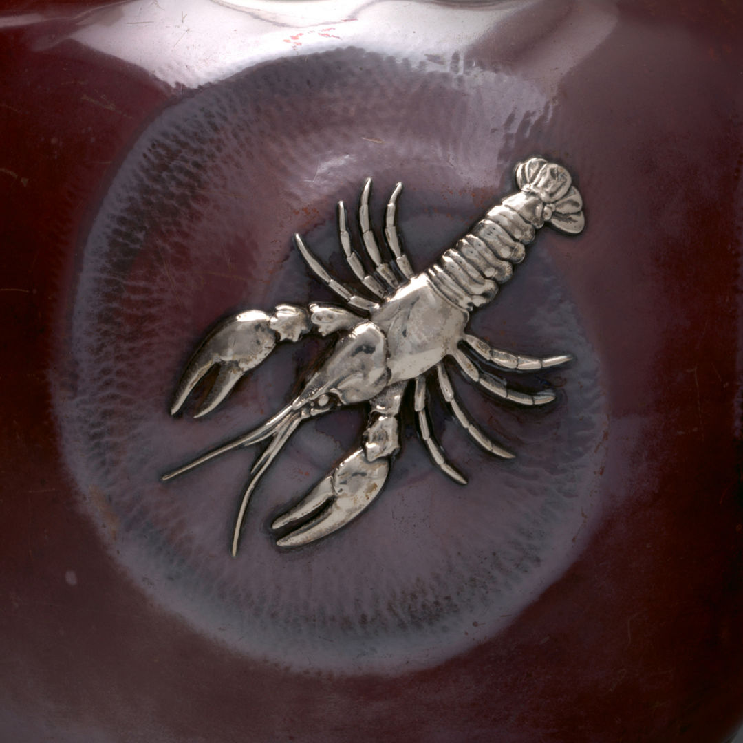 A detailed view of a patinated copper and silver chocolate pot. A lobster rendered in high-relief silver adorns the  flat circular plane of the pot’s body. 