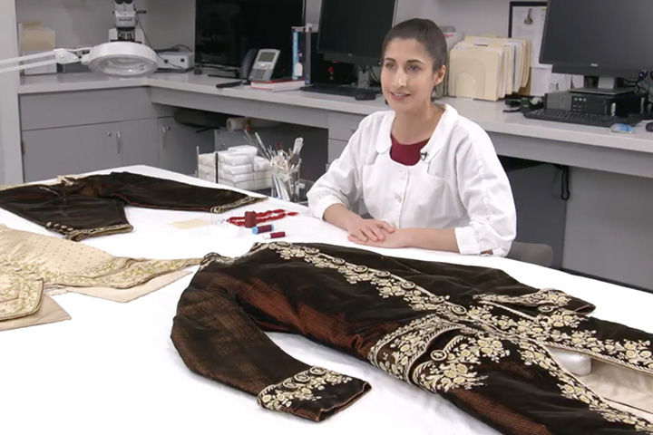 A conservationist sits at a white table. Laid out on the table in front of her are different pieces of an 18th century men's suit.