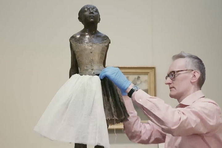 A conservationist in a gallery setting stitches the tutu on Edgar Degas's sculpture, The Little Fourteen-Year-Old Dancer.