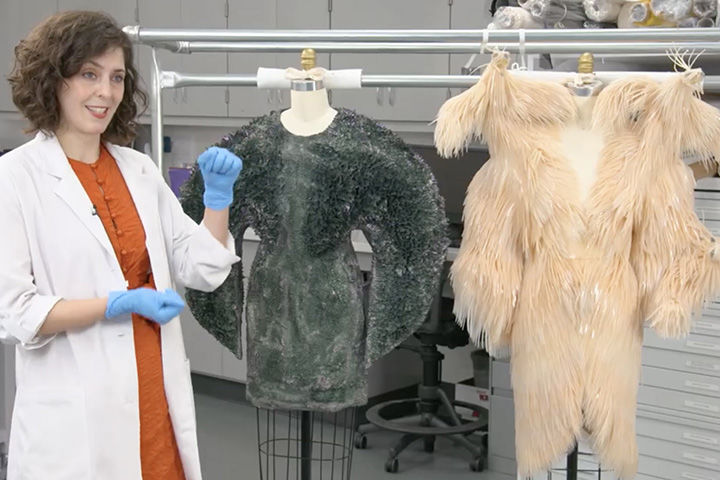 A conservationist in a white gown and blue gloves gestures towards two hanging costumes to her left: the first is a greenish-blue dress, and the second is a peach color. Both are made with unconventional materials.