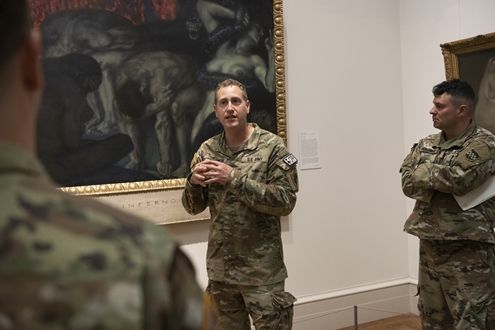 A man in military camo suit stands in front of a painting with his hands crossed, surrounded by two men turned to listen to him