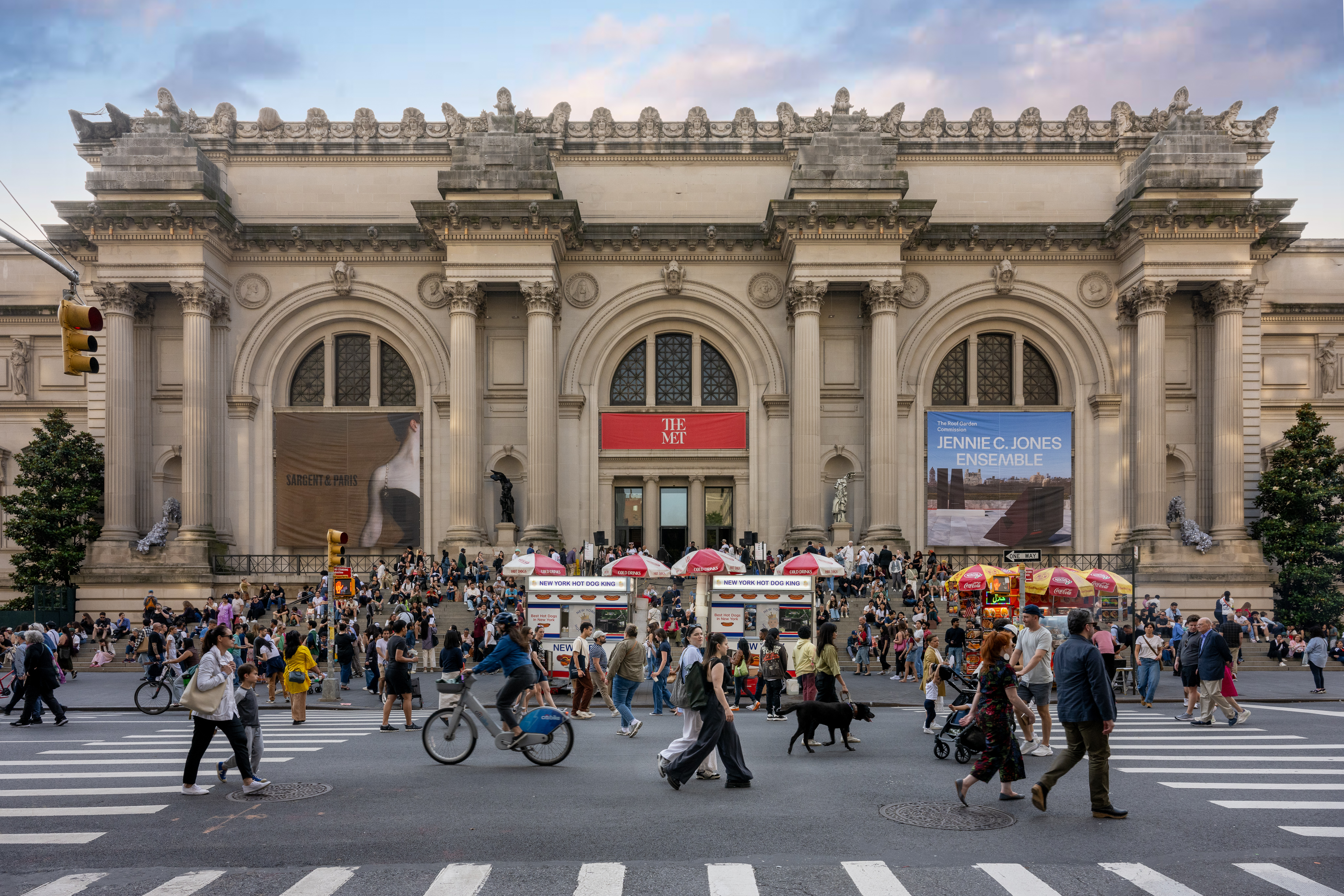 An image of the front of The Metropolitan Museum of Art on a sunny day with crowds of people walking on the sidewalk in front of the building and sitting on the front steps