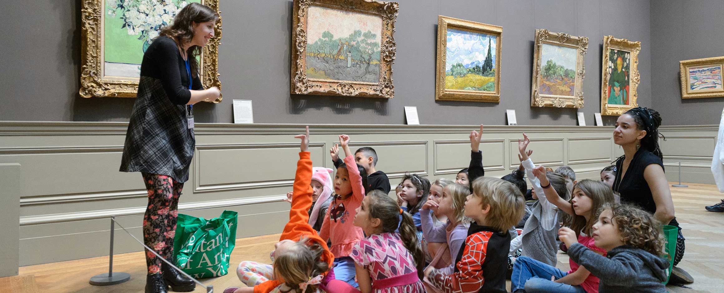 Museum educator standing in a gallery teaching a group of seated children who are raising their hands to answer a question. 