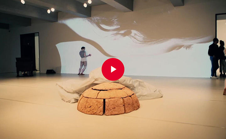 A large open room with a sculptural mound at its center; a dancer leans against a back-wall covered in a large-scale projection; a red play button appears in overlay at the center