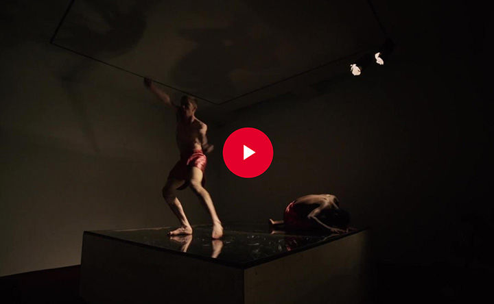 One standing dancer and one crouching dancer appears atop a dark square podium in a dark room; a red play button appears in overlay at the center