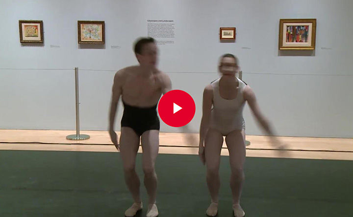 Two dancers appear side-by-side in blurred motion as they stand with bent knees as if ready to jump; they appear in front of a gallery wall of small framed artworks; a red play button appears in overlay at the center