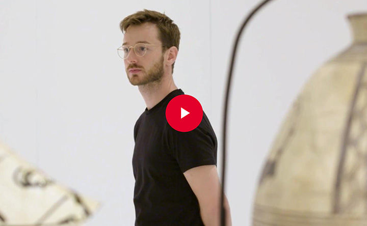 The artist Oliver Beer stands amongst vessels in a gallery space; a red play button appears in overlay at the center