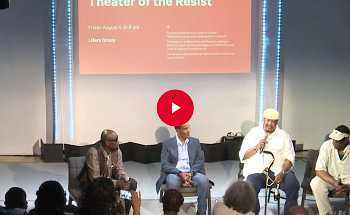 Four seated panel speakers engage in conversation on stage in front of a seated audience; a red play button appears in overlay at the center