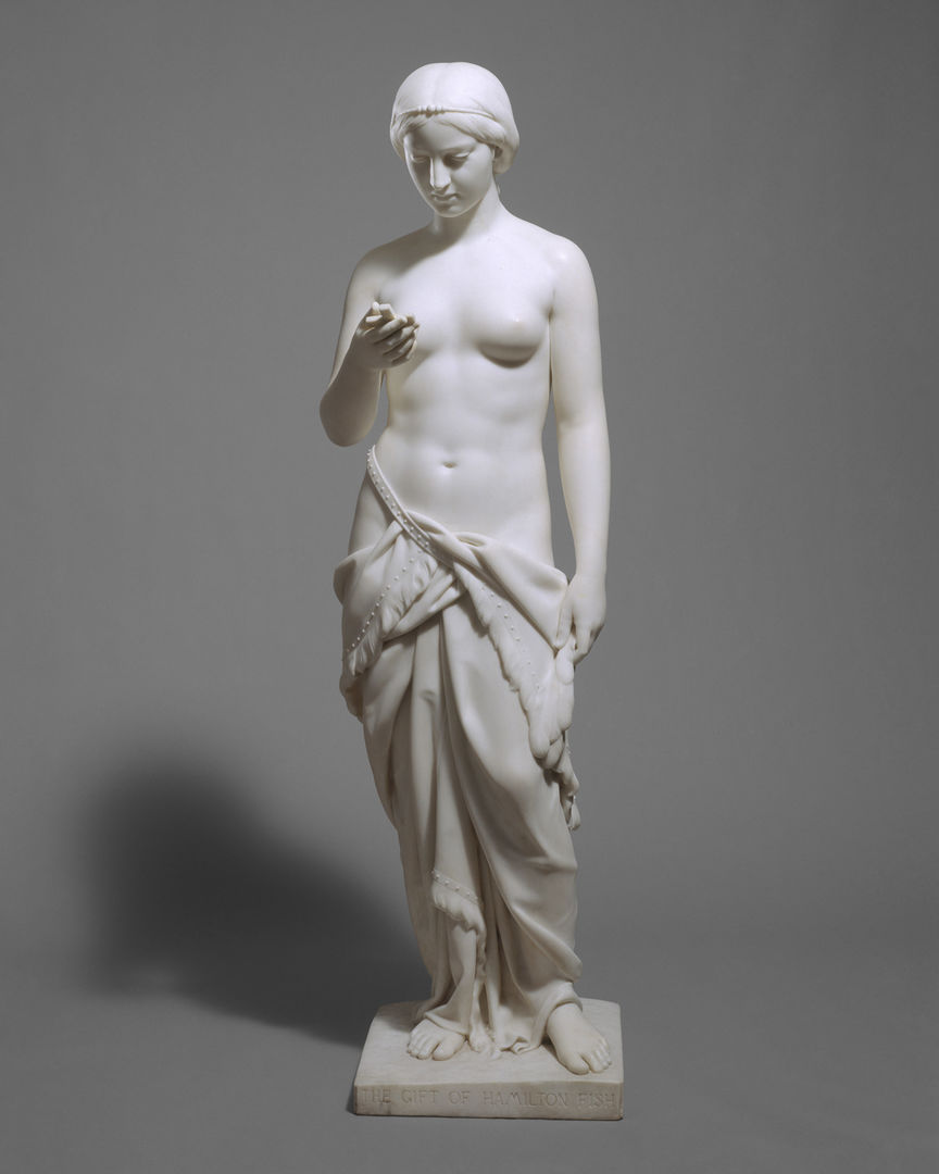 A white marble sculpture of a woman wearing a skirt, with no top, holding a crucifix in her hand