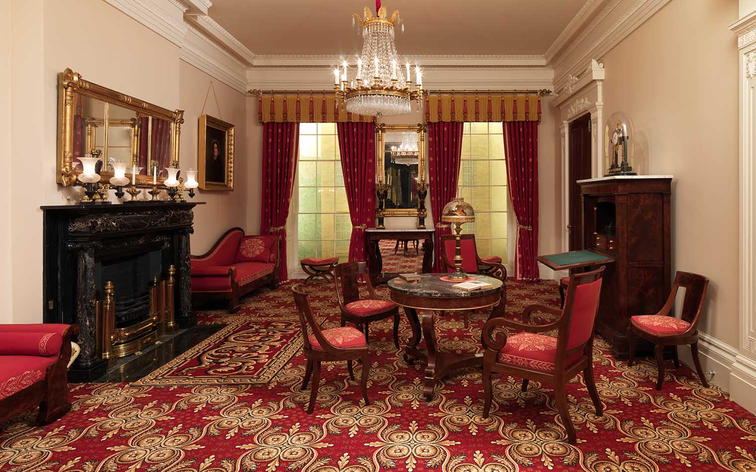 A wide view of the Greek Revival Parlor. A fireplace and mantel, table with four chairs, chandelier, and secretary are seen