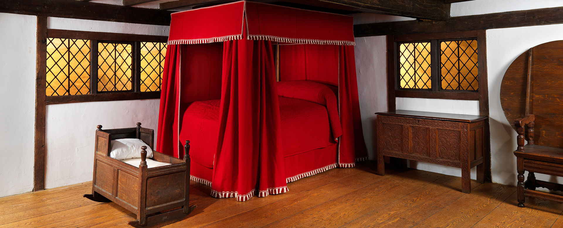 An interior image of the Hart Room showing exposed oak timbers, small casement windows, and wooden furniture including a crib and a bedstead with red curtains