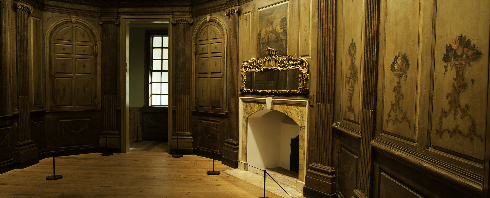 A cropped photograph of the Marmion room installed in the Met higlighting the fireplace, looking glass, and decorative wall panels.