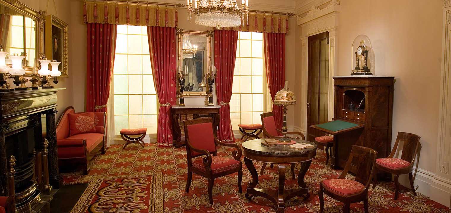 A wide view of the Greek Revival Parlor. A fireplace and mantel, table with four chairs, chandelier, and secretary are seen