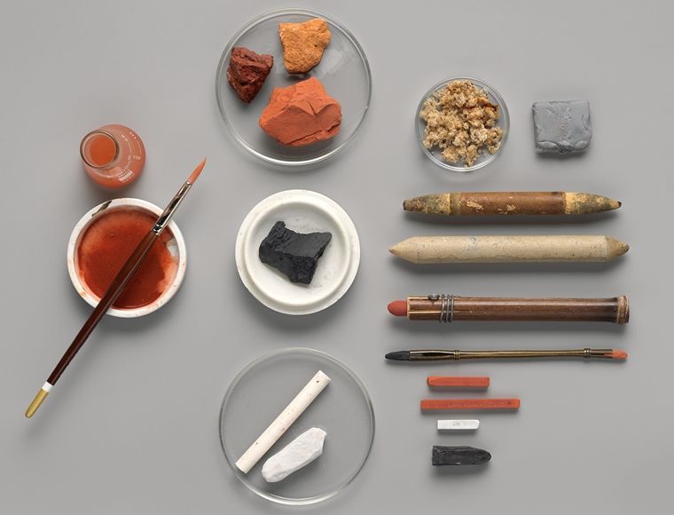 An artful arrangement of specialized tools for drawing with chalk, including multiple sticks of chalks, chalk-holders, and a small cup of water and a brush.