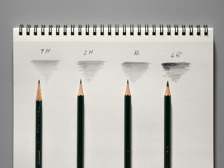 An arrangement of pencils with different hardness ratings beneath scribbles from the pencil below, displaying the darkness of each kind.