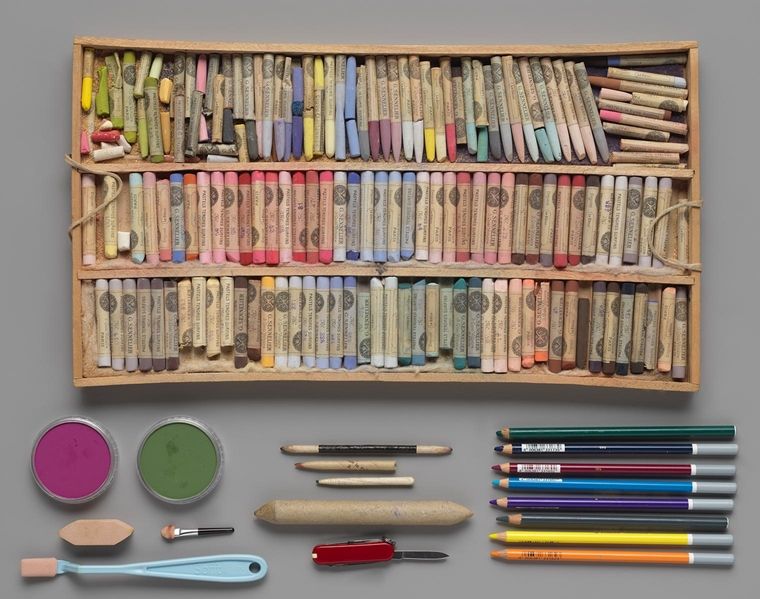 A wooden box of dozens of colorful pastel pencils, above an arrangement of other drawing implements.