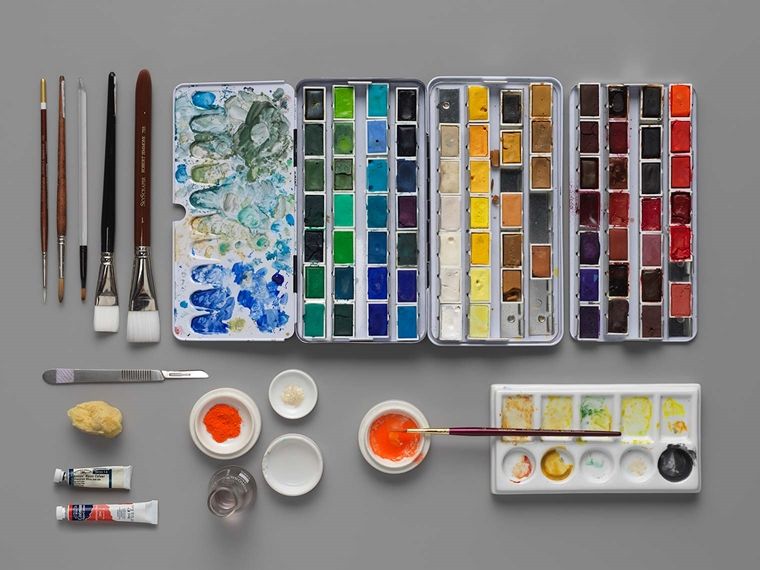 A neat arrangement of brushes, knives, watercolor pans, and other tools on a grey tabletop.