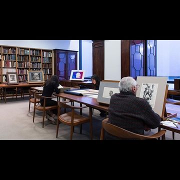 Three people sat at a long table looking at various drawings and prints on easels or flat on the table.