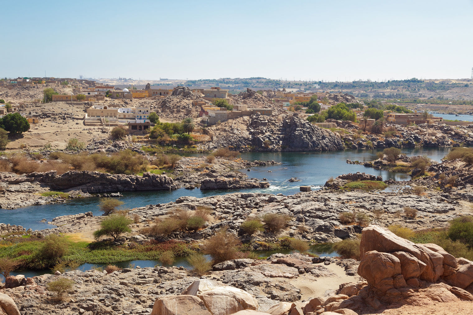 An image showing a few buildings along a shallow river in the desert. The river is the First Cataract at Aswan in Lower Nubia (modern Egypt).