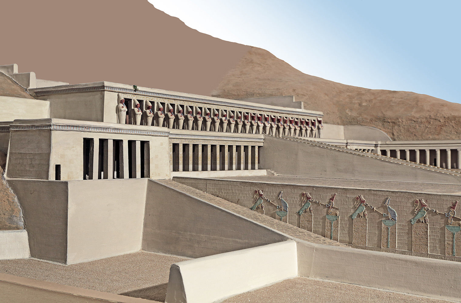 Terraced temple from the side, in a sandy landscape. On the visible wall are rectangles with crowned falcons and cobras on top, in green and red.