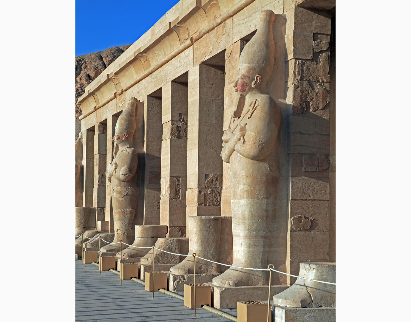 Color photograph showing the facade of a temple, with several pilasters and two complete mummiform statues wearing tall crowns visible.