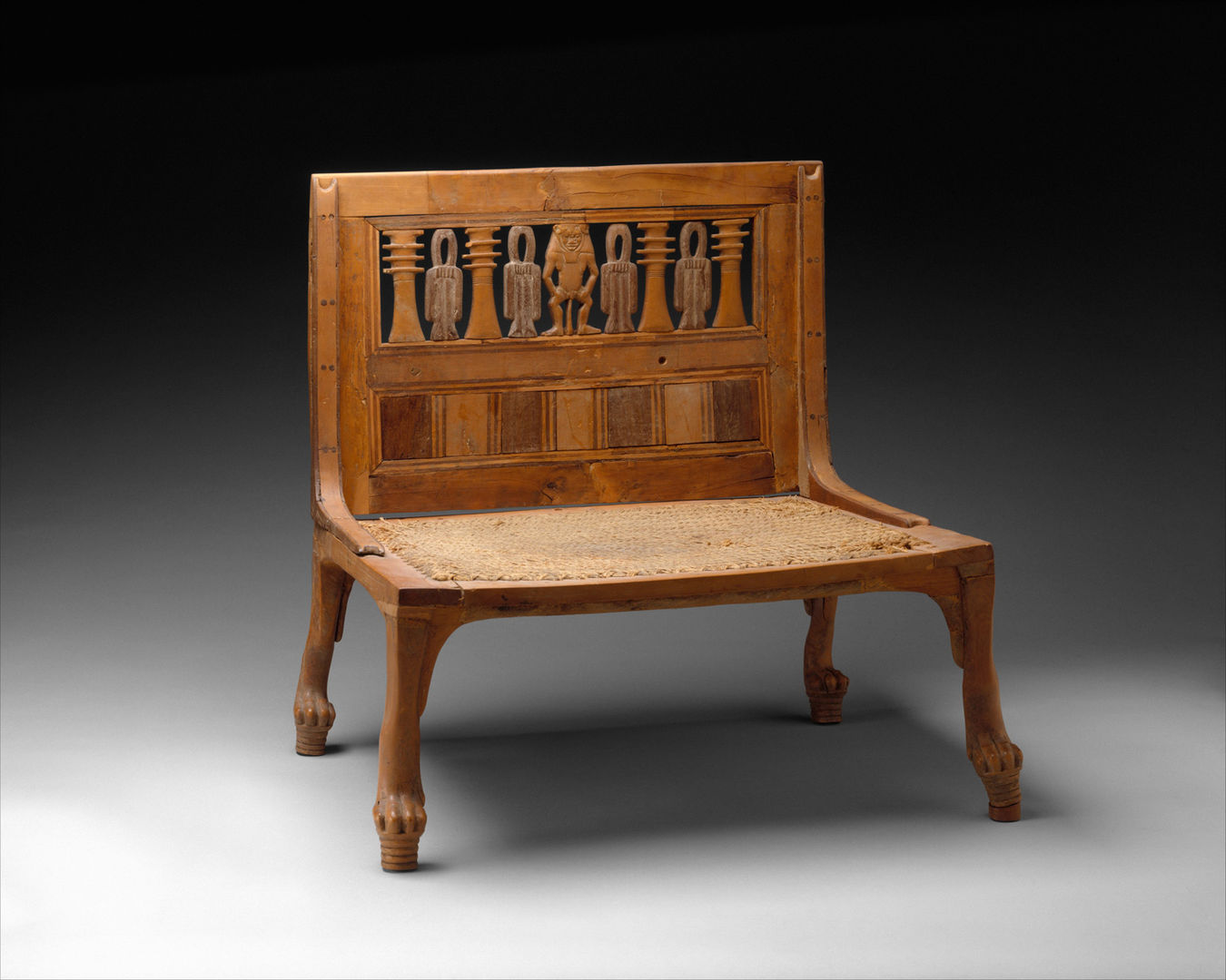 A wide wooden chair with a woven seat and lion-paw legs. The back is decorated with an upright figure with a leonine male and alternating djed pillar and tyet knot amulets.
