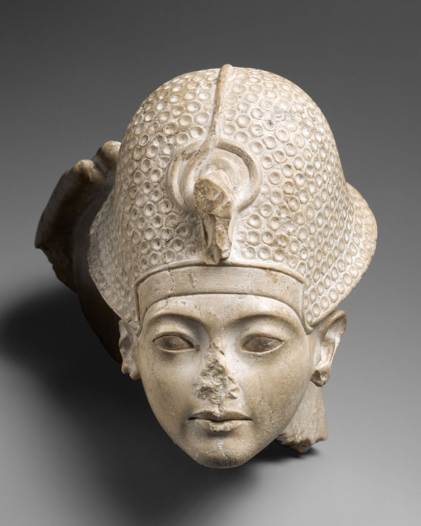 The head in white stone of a young boy wearing a helmet covered with small circles. The hand of a larger figure is visible behind the helmet on viewer left.