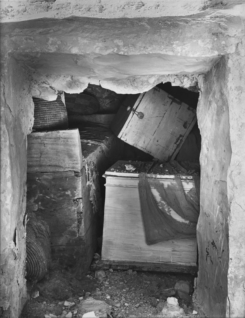 View through a rectangular doorway into a rock-cut chamber, with boxes, baskets, and a coffin visible and one tattered cloth draped over a box.
