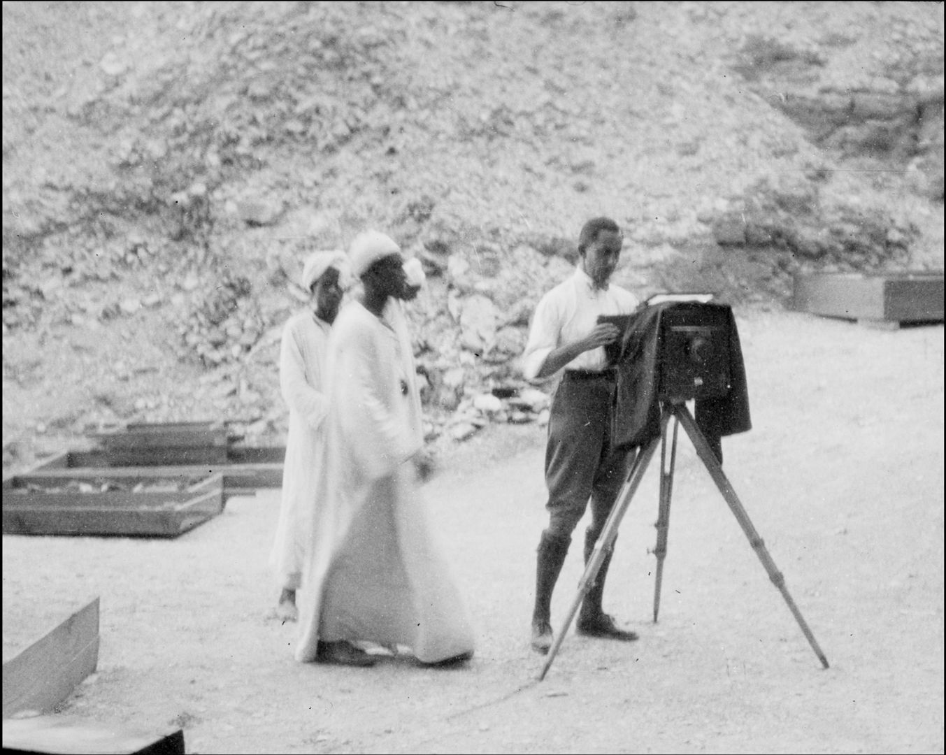 Two men in floor-length white garments stand to the left. A man in trousers and a white shirt stands to the right behind an antique camera on a tripod.