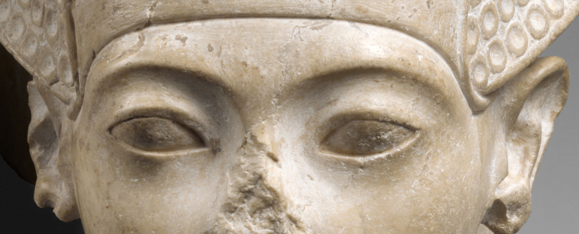 A close-up of a face, focused on the eyes, which are almond-shaped. Part of the area of the nose, which was broken away,  is visible, Above the eyes is the band of a helmet-like crown.